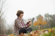 Взрослая женщина, использующая цифровые планшеты, сидит на земле в осеннем лесу . — стоковое фото