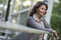 Cheerful woman in grey jacket leaning on railing and smiling in city. — Stock Photo