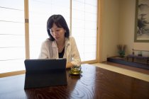 Mujer adulta sentada a la mesa con computadora portátil y taza de té verde en el interior . - foto de stock