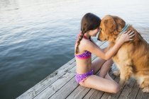 Pre-adolescent girl in swimwear with golden retriever dog sitting on jetty. — Stock Photo