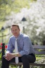 Uomo d'affari in camicia e cravatta seduto sulla panchina del parco sotto l'albero con fiore . — Foto stock