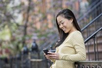 Asian woman leaning on railing on city street and using smartphone. — Stock Photo