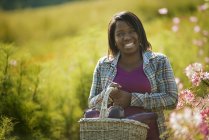 Mulher sorrindo e segurando cesta de berinjelas em prado florido na fazenda orgânica — Fotografia de Stock