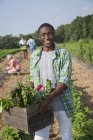 Teenage boy holding wooden box of fresh vegetables harvested from field with people in background. — Stock Photo
