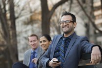 Businessman holding smartphone with colleagues relaxing on bench in city park. — Stock Photo
