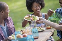 Young friends sharing plates with food at picnic table in countryside garden. — Stock Photo