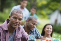 Jeune homme regardant à la caméra avec des amis à la table de pique-nique dans le jardin de campagne . — Photo de stock
