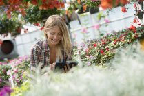Junge Frau untersucht Blumen mit digitalem Tablet in Gärtnerei. — Stockfoto