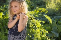 Mädchen im Grundschulalter mit lockigem Haar steht im sonnigen Garten und hält frisch gepflückte Möhren in der Hand. — Stockfoto