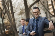 Businessman holding smartphone with colleagues relaxing on bench in city park. — Stock Photo
