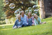 Family sitting on countryside lawn surrounded by bubbles in air. — Stock Photo