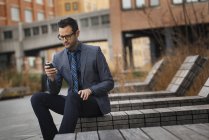Mann in offizieller Kleidung sitzt auf Bank und checkt Smartphone in der Stadt. — Stockfoto