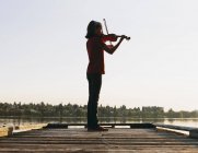 Pre-adolescent female violinist playing violin at dawn on wooden pier at lake. — Stock Photo