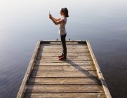 Pre-adolescent girl holding digital tablet while standing on wooden dock over water. — Stock Photo