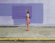 Pre-adolescent girl holding violin and bow in hands on street against purple wall background. — Stock Photo