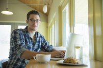 Young man sitting at cafe table with laptop and looking in camera. — Stock Photo