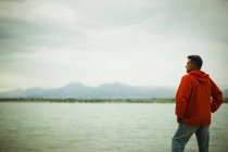 Hombre maduro en chaqueta roja mirando a la vista en el campo orilla del lago . - foto de stock