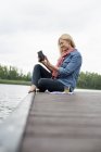Vista lateral da mulher sentada no molhe pelo lago e usando o tablet digital . — Fotografia de Stock