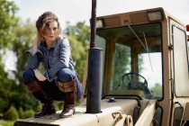 Giovane donna in giacca di jeans e stivali accovacciata sul cofano del trattore . — Foto stock