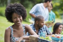 Mujer joven sonriendo en cámara con amigos en la mesa de picnic en el jardín del campo . - foto de stock