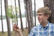 Pre-adolescent boy standing among trees on shore of lake and holding twig. — Stock Photo