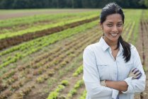 Woman standing with arms crossed in front of rows of green vegetable plants on organic farm. — Stock Photo