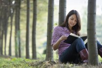 Donna seduta a leggere libro sotto gli alberi nel bosco . — Foto stock