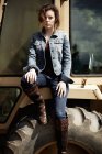 Young woman in denim jacket and boots sitting on hood of tractor. — Stock Photo