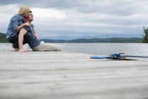 Woman hugging man while sitting together on jetty by lake. — Stock Photo