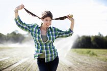 Mujer joven con camisa verde a cuadros sosteniendo trenzas mientras está de pie en el campo con aspersores . - foto de stock