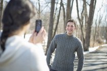 Woman taking picture of young man with smartphone in wintry woods. — Stock Photo