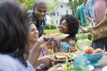 Young friends sharing plates with food at picnic table in countryside garden. — Stock Photo