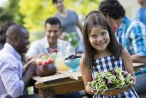 Elementary age girl holding bowl of salad with adults around table in garden. — Stock Photo