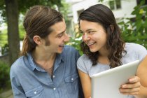 Young couple sitting side by side, looking and each other and holding digital tablet. — Stock Photo