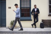 Man in full suit leaning against wall with Asian man walking past and checking phone. — Stock Photo