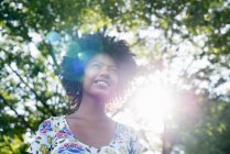 Young woman in flowered dress smiling and looking up in sunny forest. — Stock Photo