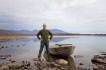 Uomo maturo in piedi sulla riva del lago accanto barca a remi . — Foto stock