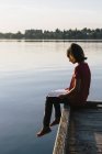 Pre-adolescent girl sitting on wooden dock and reading book. — Stock Photo