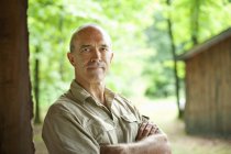 Mature man standing with arms crossed in front of wooden hut in countryside — Stock Photo