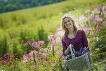 Mujer sonriendo y sosteniendo cesta de berenjenas en prado florido en granja orgánica - foto de stock