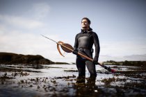 Hombre en traje de neopreno de pie en la orilla con lanza de arpón de pesca . - foto de stock