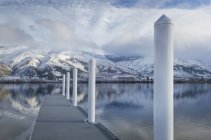 Pilastri sul molo al lago vicino alla catena montuosa innevata — Foto stock