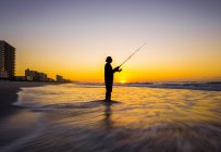Blurred view of silhouette of man fishing in waves on beach at sunset — Stock Photo