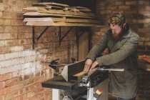 Hombre barbudo de pie en el taller, con protectores de oídos, trabajando en pieza de madera . - foto de stock