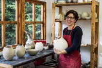 Frau in roter Schürze steht in Werkstatt, hält Keramikvase in der Hand und lächelt in die Kamera. — Stockfoto