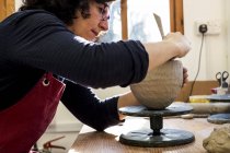 Donna in grembiule rosso seduta in laboratorio di ceramica e che lavora su vaso di argilla . — Foto stock