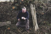 Bearded man sitting on ground next to bunch of wooden stakes, holding mug, checking mobile phone. — Stock Photo