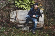 Bearded man wearing black beanie sitting on wooden bench in garden, holding blue mug, looking in camera. — Stock Photo