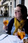 Mujer sentada a la mesa al aire libre en la ciudad de Venecia y sosteniendo un vaso de bebida alcohólica, Italia - foto de stock