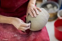 Manos de artista de cerámica en delantal rojo sentado en el taller, trabajando en jarrón de arcilla . - foto de stock